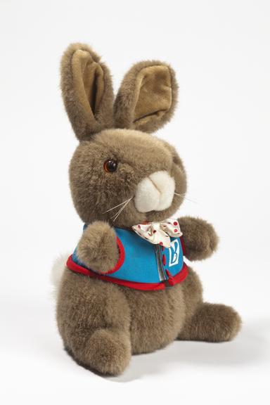 Brown plush rabbit toy seated against a white background, wearing a blue vest with red trim, a patterned white neckerchief, and a small Rabbit logo badge.