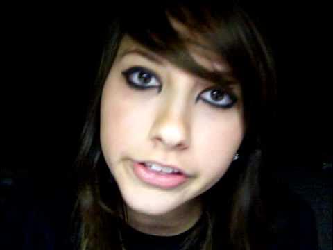 Low-resolution close-up of a young woman with dark side-swept hair and heavy eyeliner looking toward the camera against a nearly black background.