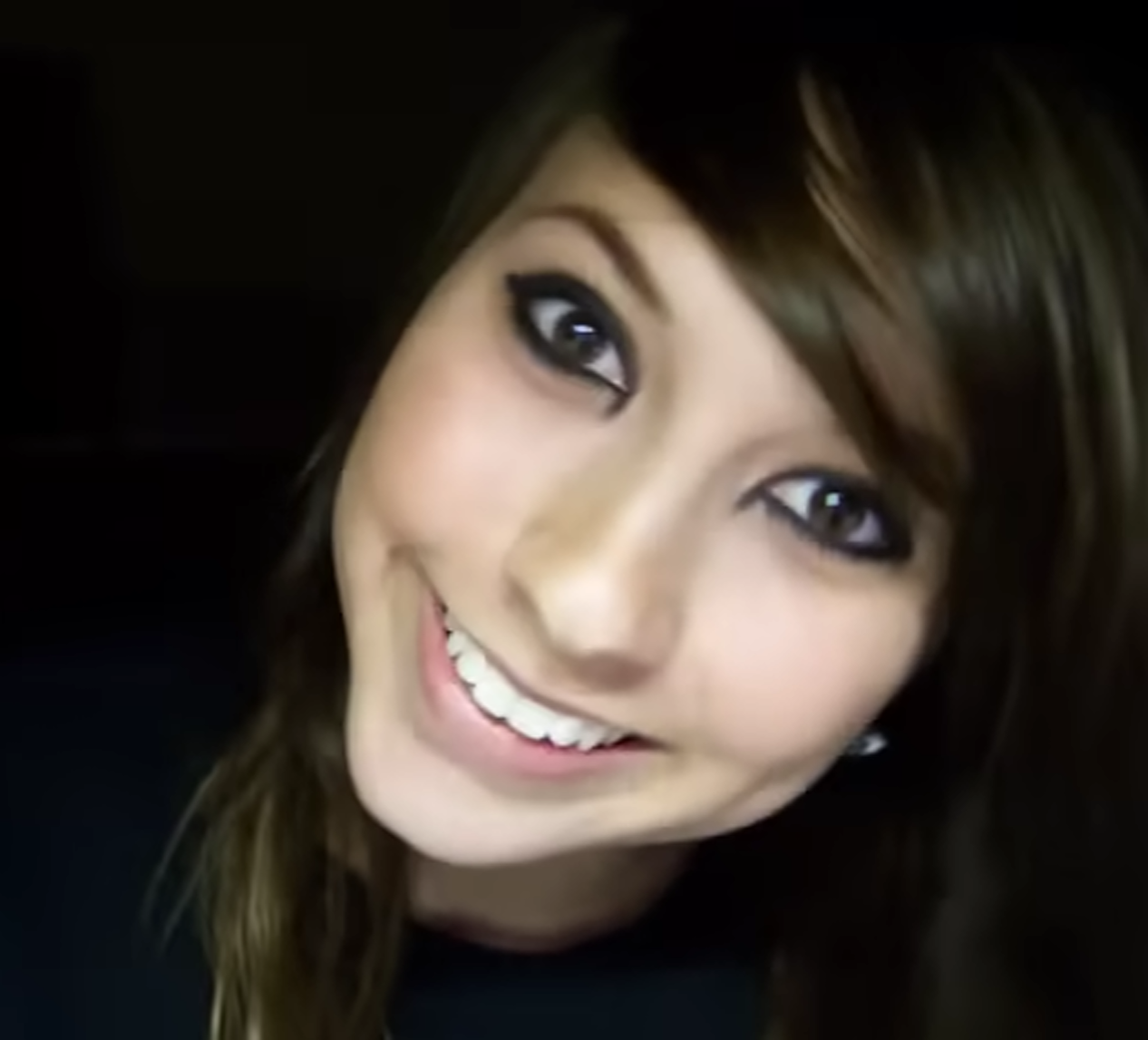 Close-up of a smiling young woman with long brown hair and heavy black eyeliner, her head tilted against a dark background.