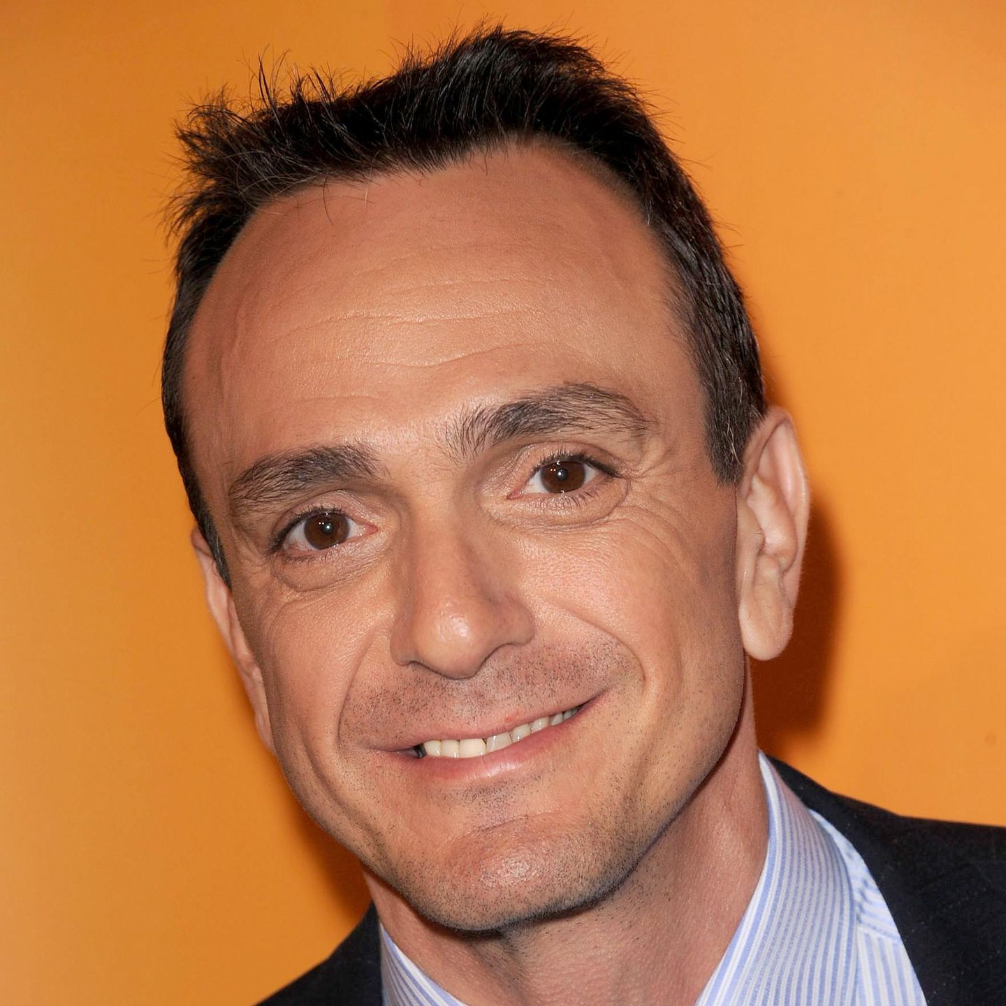 Close-up portrait of a smiling man with short dark hair wearing a striped collared shirt against an orange background.