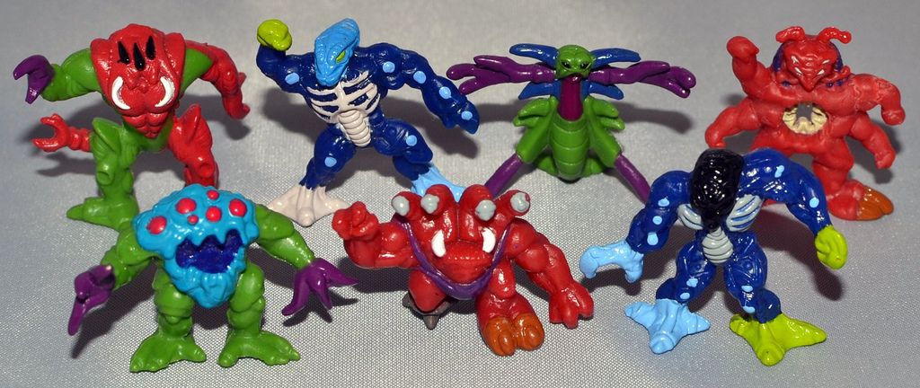 Seven small colorful alien figures arranged on a gray surface, including red, green, blue, purple, and turquoise creatures with varied monster-like bodies.