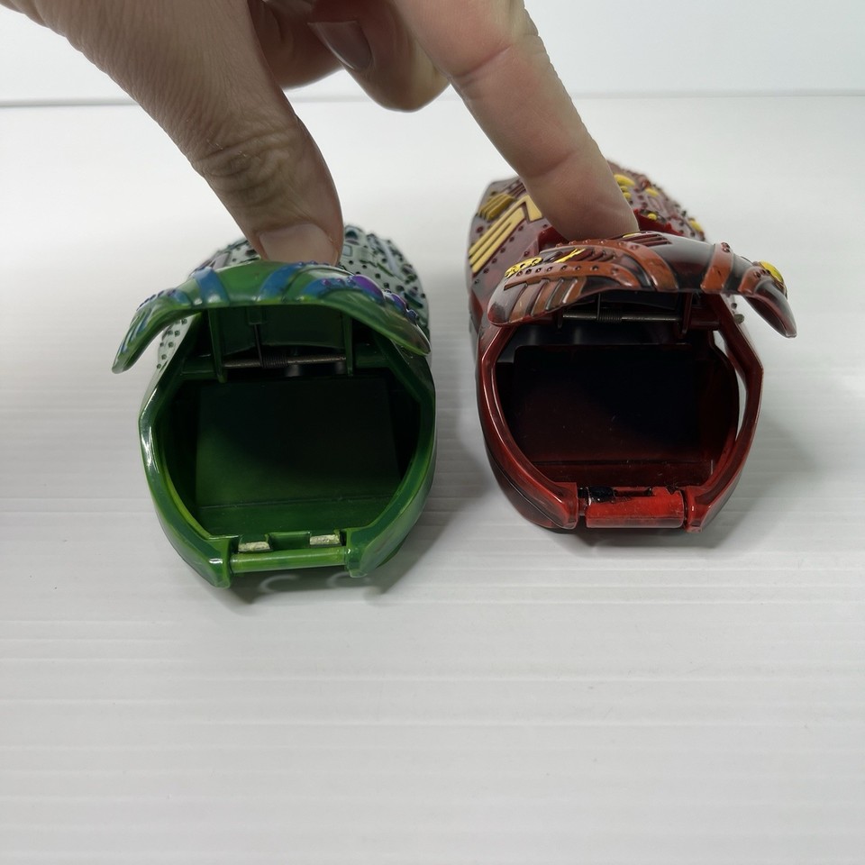 Close view of two open plastic Space Pods on a white surface, one green and blue and one red and brown, with a person’s fingers holding up the hinged top of the red pod.
