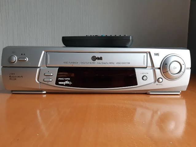 Front view of a silver VHS video cassette recorder on a wooden surface, with a black remote control resting on top. The front panel has a cassette slot, circular playback control, buttons, and a VideoPlus+ logo on the dark display area.