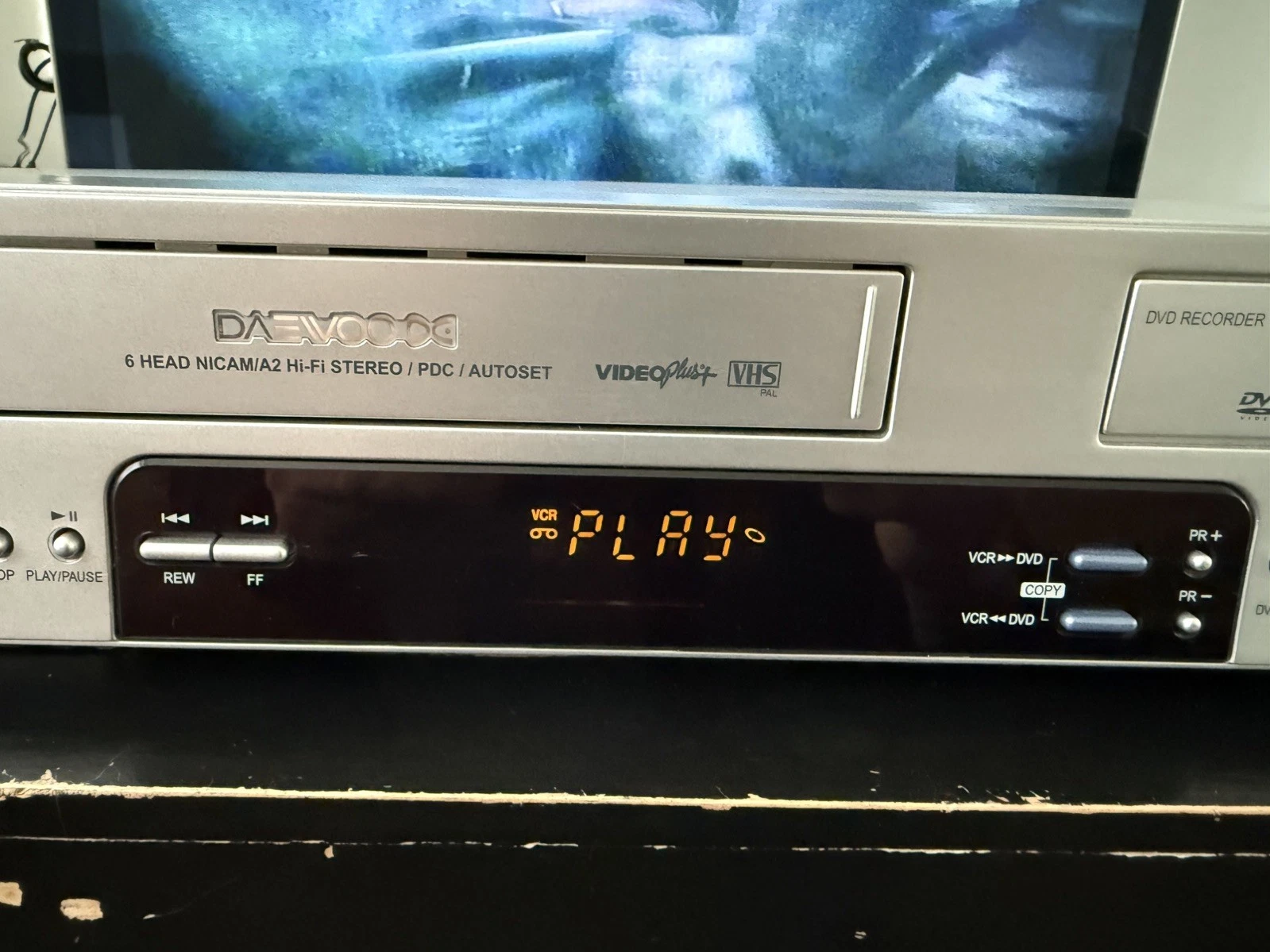 Close-up of the front of a silver Daewoo VHS/DVD recorder. Text beside the VHS cassette door includes “VideoPlus+,” and the display reads “VCR GO PLAY.”