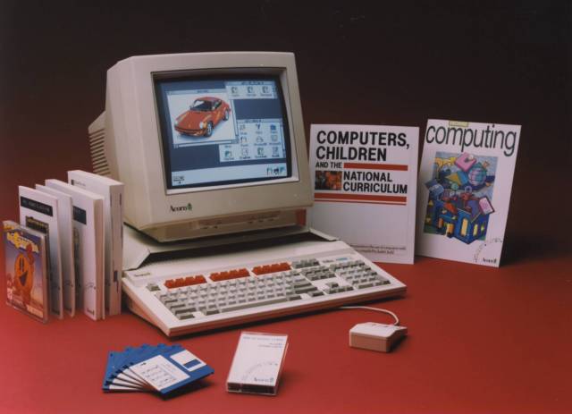 Acorn desktop computer setup on a red surface, with CRT monitor showing a car image, keyboard, mouse, floppy disks, software boxes and books including “Computers, Children and the National Curriculum.”