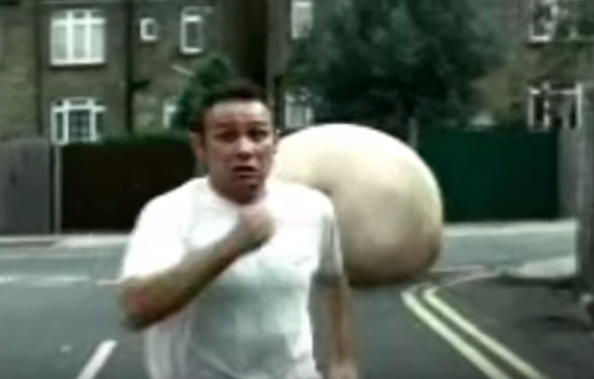 Low-resolution still of a man in a white T-shirt running toward the camera on a city street, with a large flesh-colored rounded object behind him near double yellow road lines.