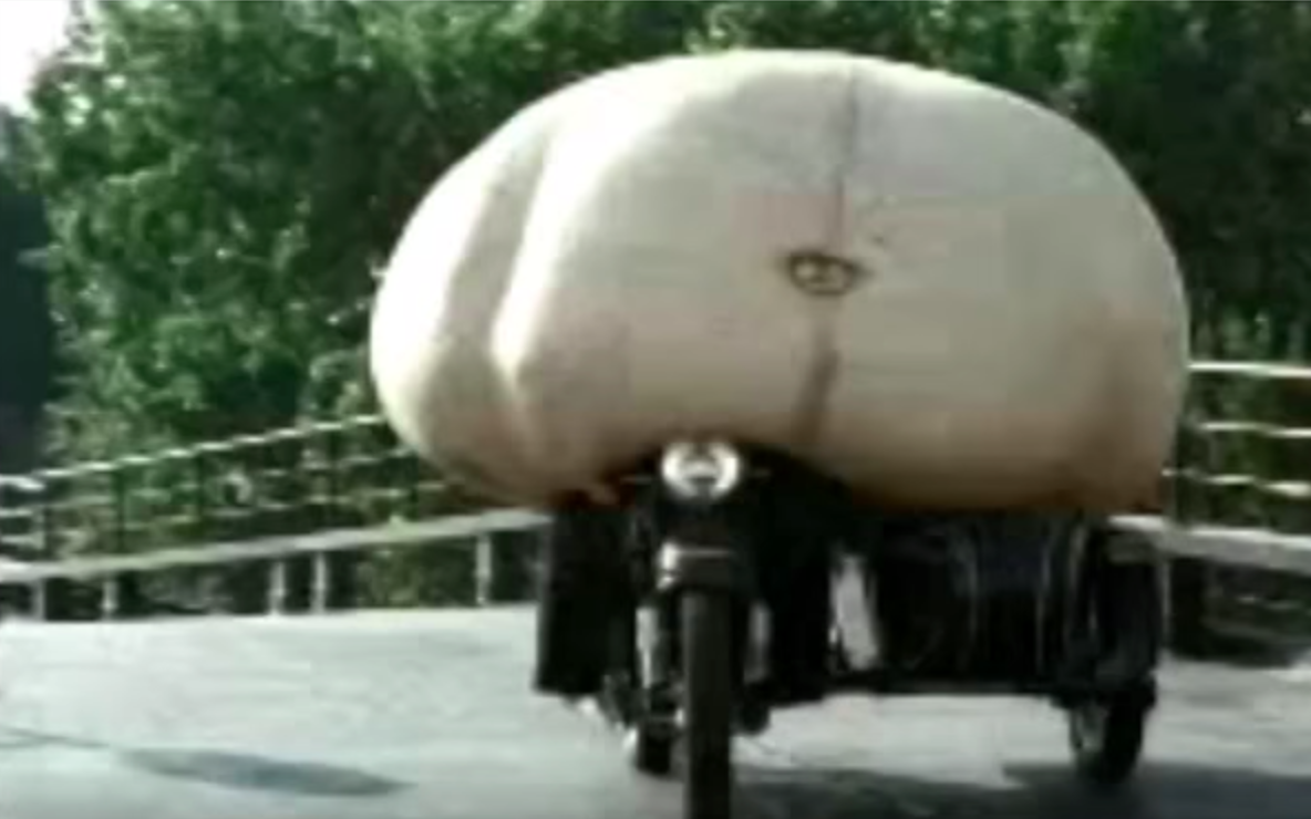 A large flesh-colored belly-shaped form with a visible navel is mounted over a dark motorcycle or motorized vehicle on a paved path beside trees and a metal railing.
