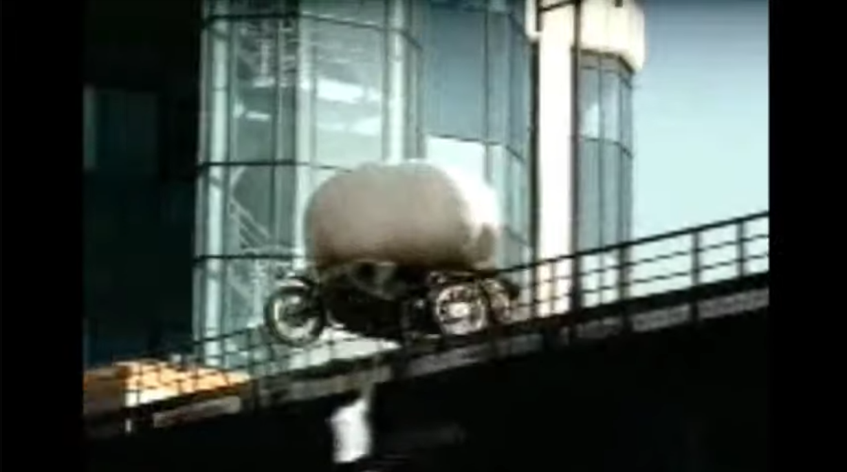 A large flesh-colored rounded object rides above a dark wheeled vehicle on an elevated roadway beside a glass-fronted building.