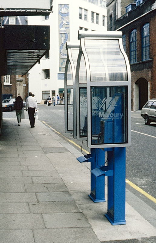 Two blue freestanding public telephone units with curved silver-and-glass hoods stand on a city pavement beside a road; the nearer unit displays a Mercury Communications logo, with pedestrians and office buildings behind.