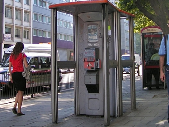 A weathered grey public payphone unit stands inside a narrow glass-sided street shelter beside a pavement; a woman walks past, cars are parked behind, and a red BT-branded telephone shelter is partly visible in the background.