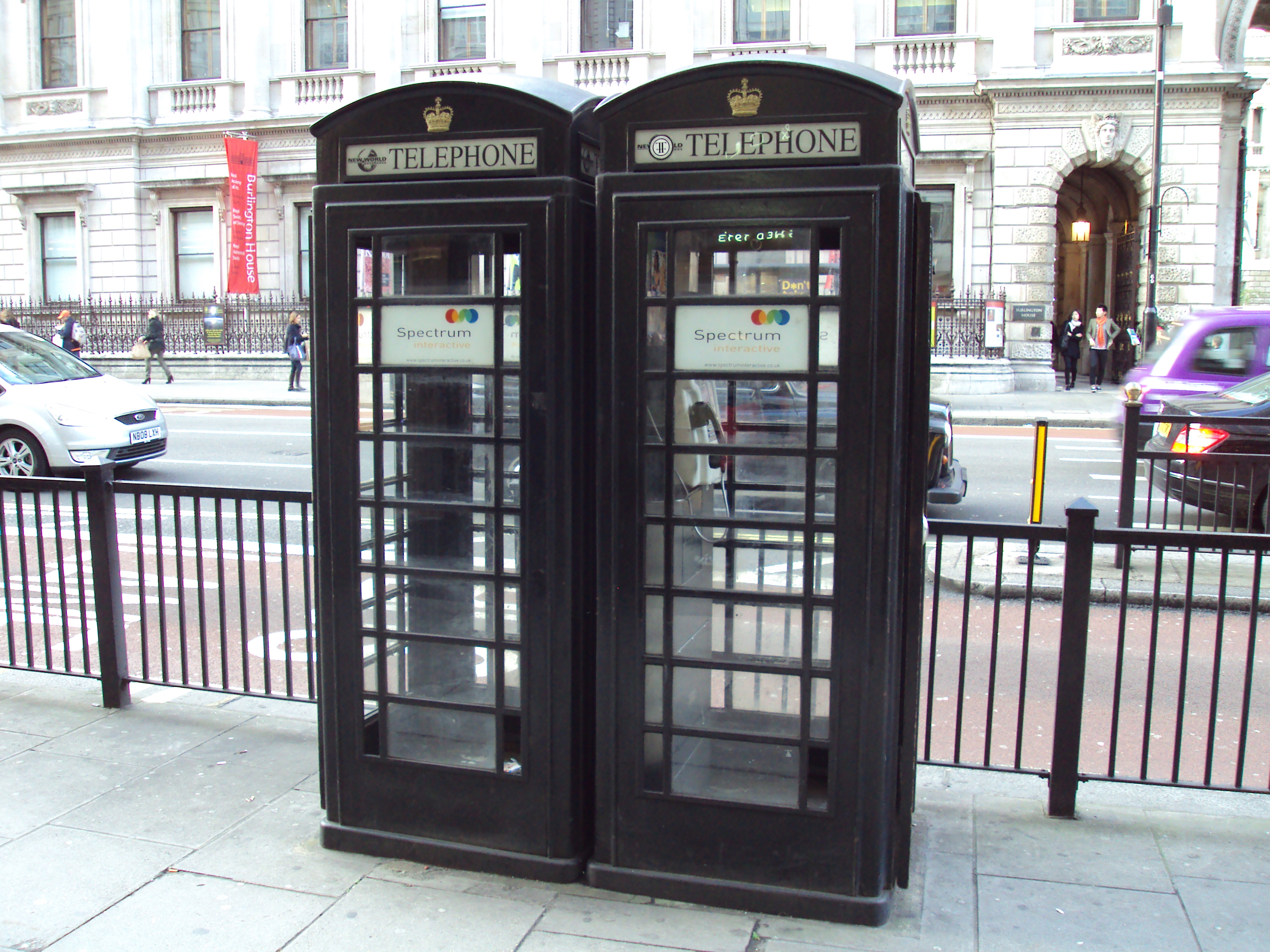 Two black glass telephone kiosks stand on a city pavement, each with a sign reading "NEW WORLD TELEPHONE" and Spectrum Interactive advertisements in the lower panels; cars and pedestrians pass in front of a stone building.