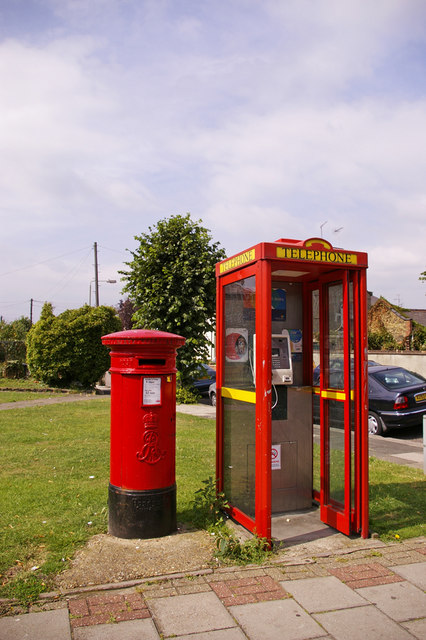 A red glass telephone kiosk with a yellow "TELEPHONE" sign stands beside a red cylindrical post box on a grassy roadside verge, with parked cars and houses behind.
