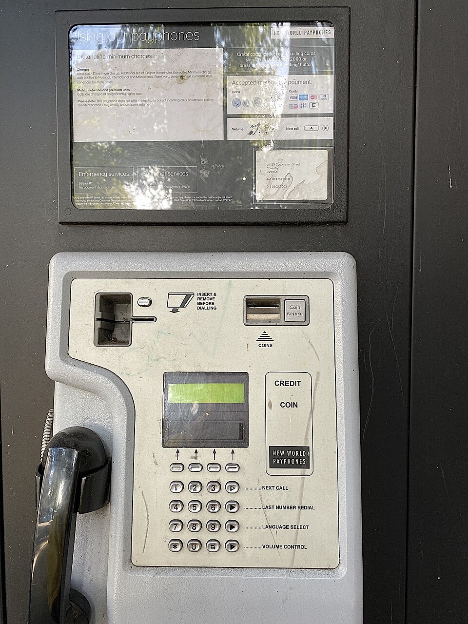 Close-up of a New World Payphones public telephone with a black handset, card slot, coin slot, display, keypad, payment instructions, and emergency-services information panel.