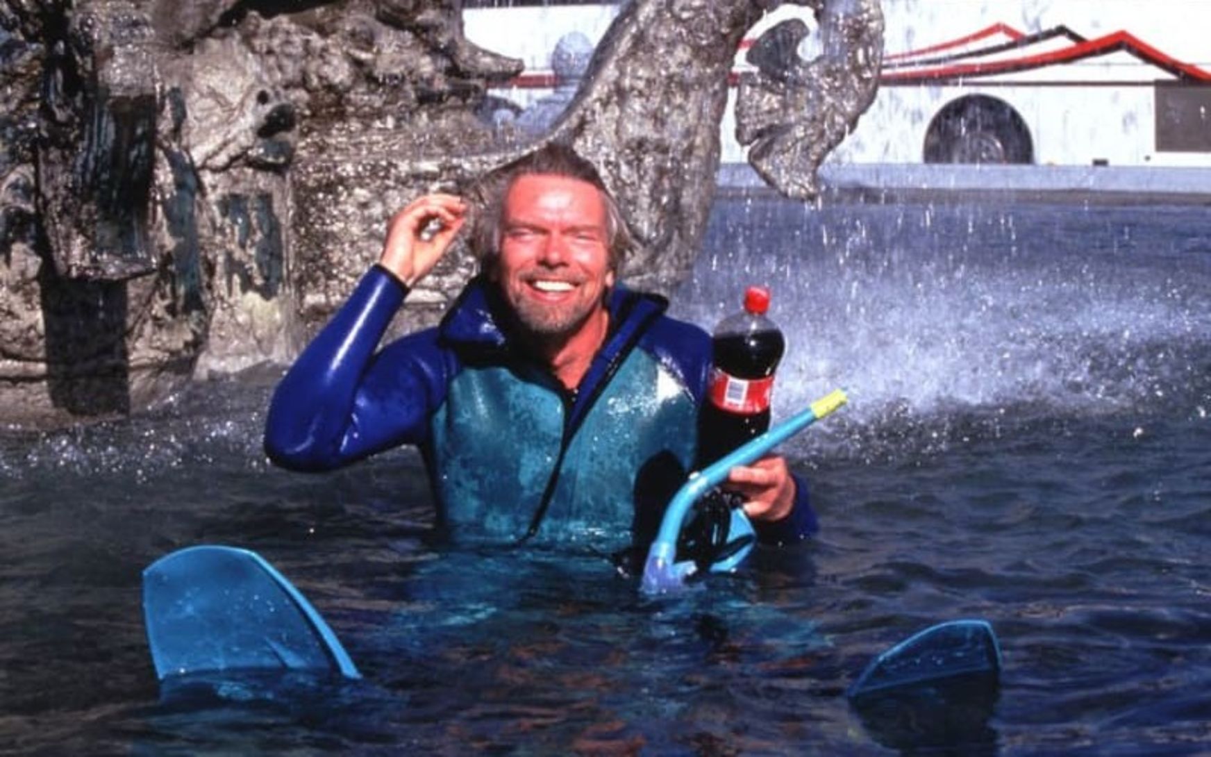 A smiling man in a blue wetsuit stands chest-deep in a fountain, holding a dark bottle with a red label beside a snorkel mask and blue swim fins.