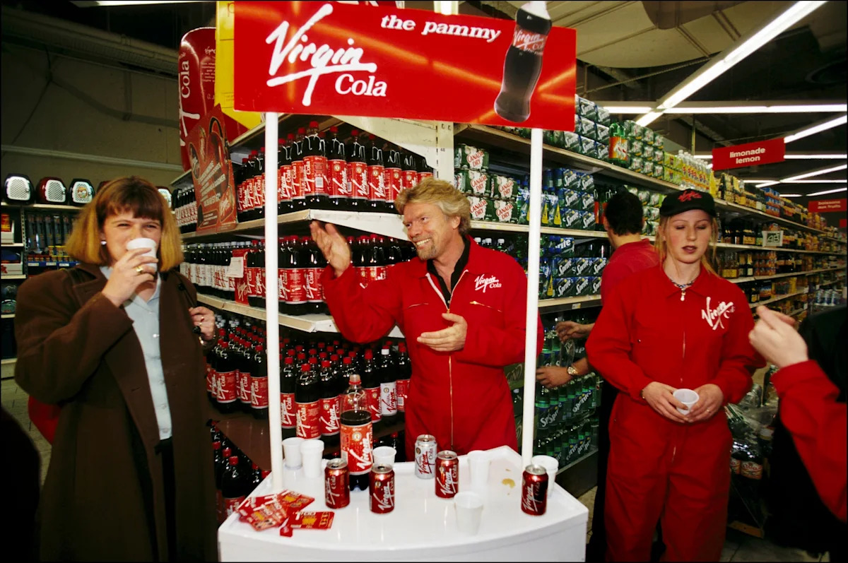 Inside a supermarket, a man and two women in red Virgin Cola-branded clothing offer small paper cups from a tasting stand beneath a red sign reading “Virgin Cola the pammy”; shelves behind them are stocked with soft drinks and Virgin Cola bottles.