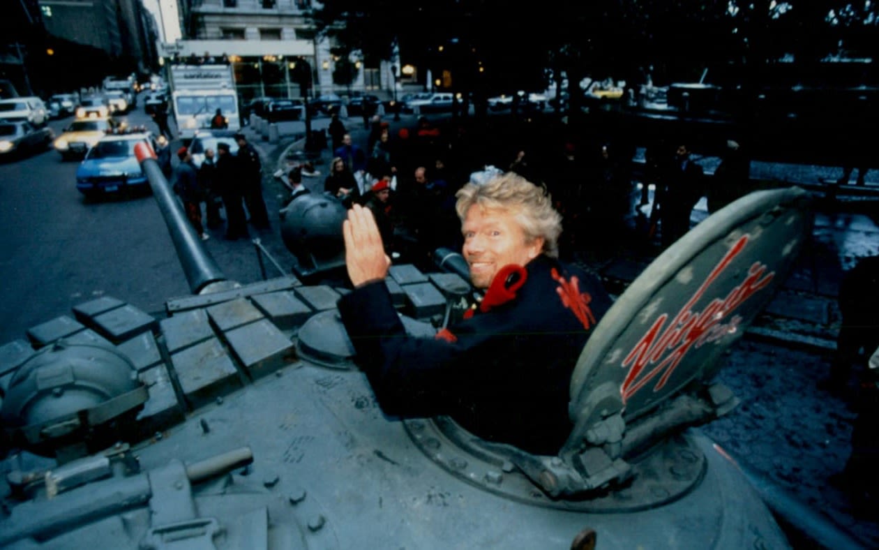 A blond man waves from the open hatch of a grey tank on a city street; a red Virgin logo is painted on the hatch cover, with traffic and onlookers behind him.