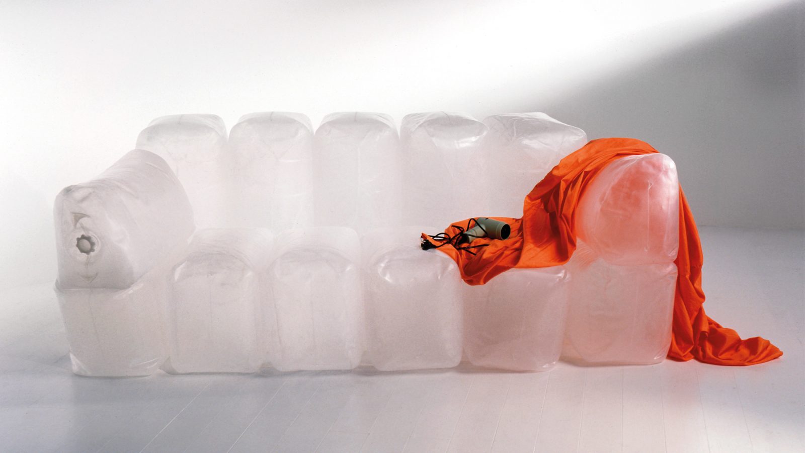 Studio photograph of a transparent inflatable sofa with a segmented back and armrests; an orange fabric cover is draped over the right arm beside a small electric pump and black cord.