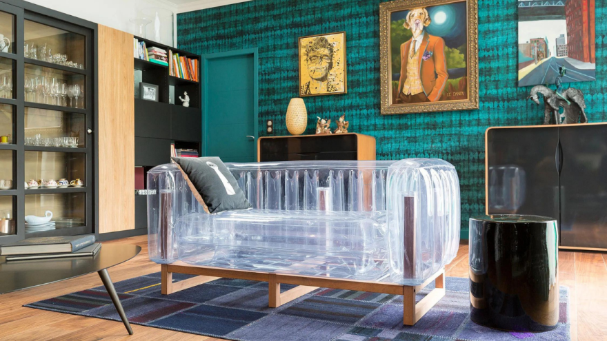 A transparent inflatable sofa on a wooden frame stands on a patterned rug in a furnished room with teal patterned walls, framed paintings and dark cabinets.