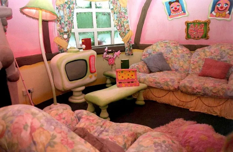Pink living-room interior with floral sofas, a green floor lamp, a small rounded cream television, a low green table holding a pink spotted box, a window with patterned curtains, and cartoon-style pictures on the walls.