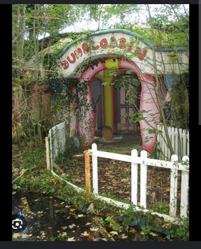 An overgrown entrance to a small pink-and-white themed structure, with a curved painted sign partly obscured by foliage, a white wooden gate, and fallen leaves on the path.