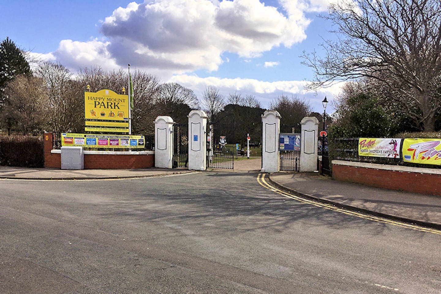 Street-facing entrance to Happy Mount Park, with white gate pillars, black gates, a yellow park sign, roadside banners and leafless trees beneath a blue sky with clouds.