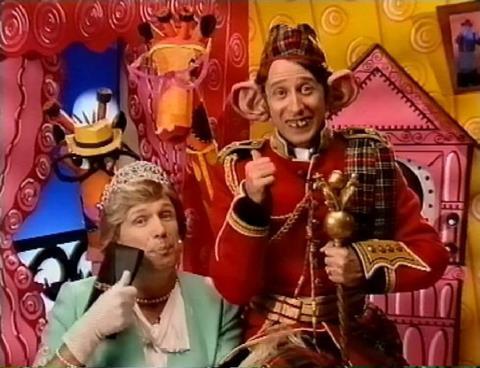 Two costumed performers pose in a brightly colored studio set decorated with spirals and cartoon-like props; the seated performer holds a dark rectangular object, while the standing performer holds a small gold statuette.