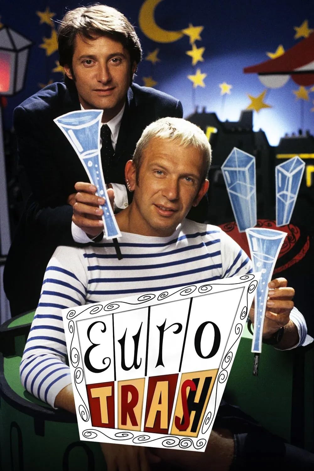 Two men pose on a brightly colored studio set beneath a crescent moon and stars, holding illustrated blue champagne flutes behind a large sign reading “Euro Trash.”