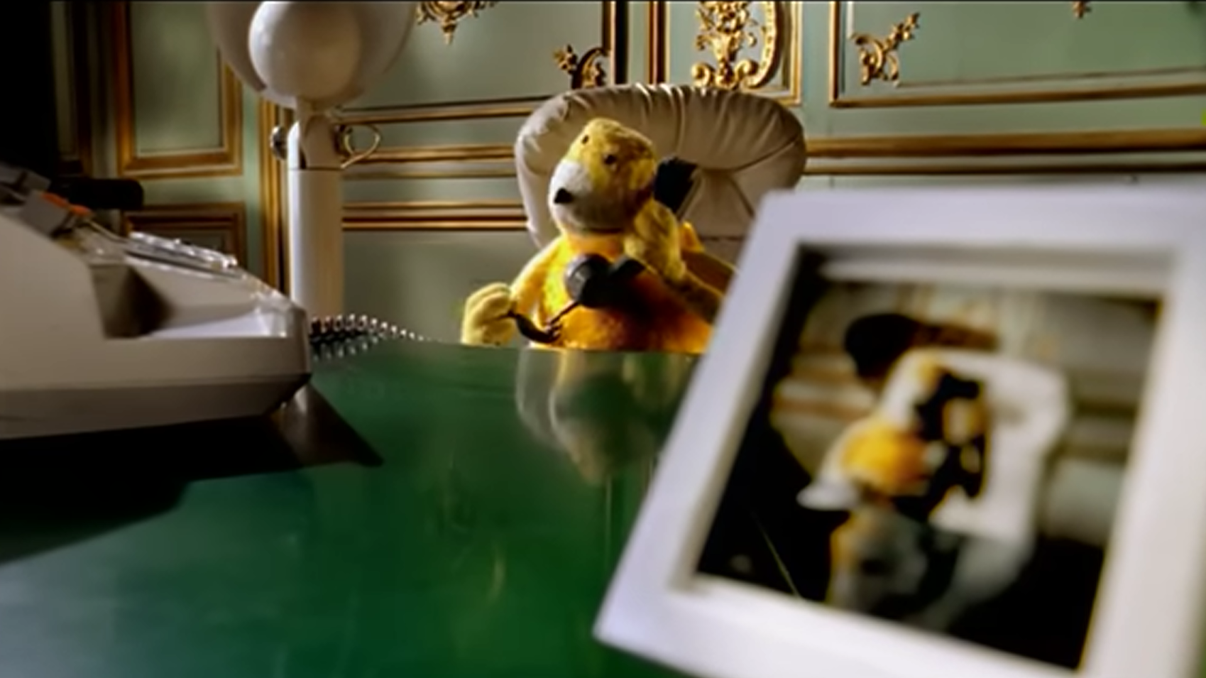 Yellow fuzzy puppet seated behind a glossy green desk, holding a black telephone receiver beside its head; a typewriter sits at left and a blurred framed photograph fills the foreground of an ornate green-and-gold room.