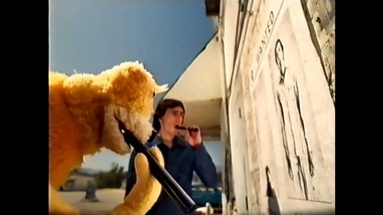 Flat Eric holds a long black gun beside a weathered white vehicle while a dark-haired man in a blue shirt drinks from a bottle in the background.