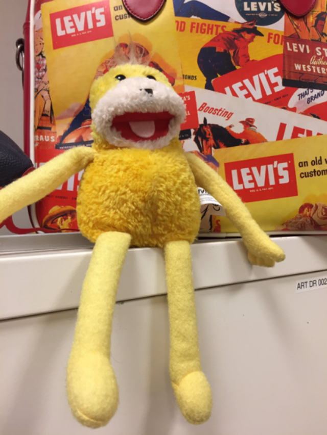 A yellow Flat Eric plush toy with long arms and legs sits on a white cabinet in front of a suitcase covered with colorful vintage-style Levi’s labels and illustrations.
