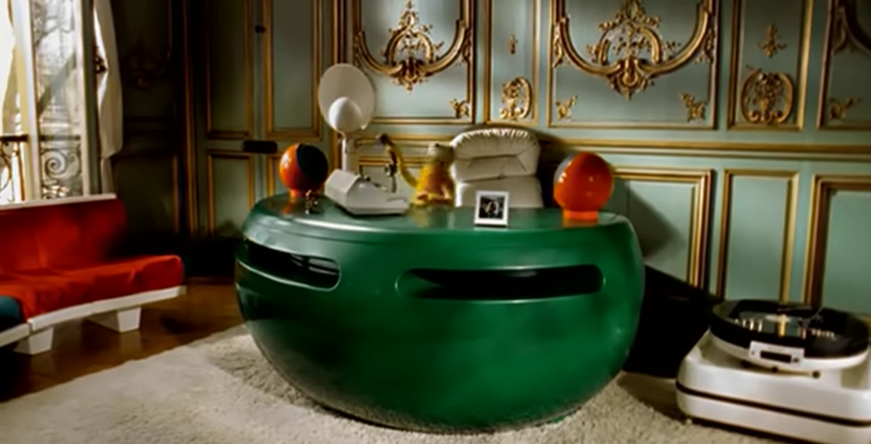 A small yellow fuzzy puppet sits behind a large curved green desk in an ornate pale-green and gold room, surrounded by a white chair, lamps, a framed photograph and audio equipment.