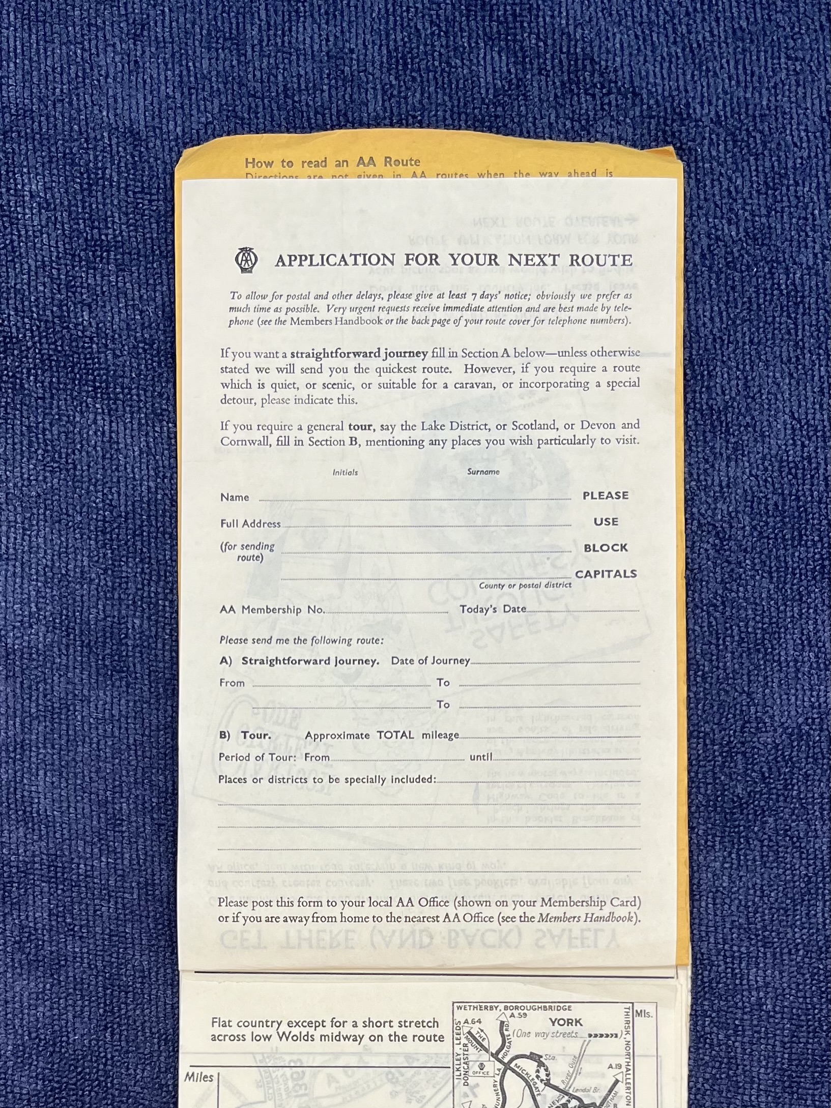 An unused AA “Application for Your Next Route” form lies over a yellow route document on dark blue fabric. It includes blank fields for member details, journey dates, destinations, tour mileage and requested stops, with part of a printed route map visible beneath.