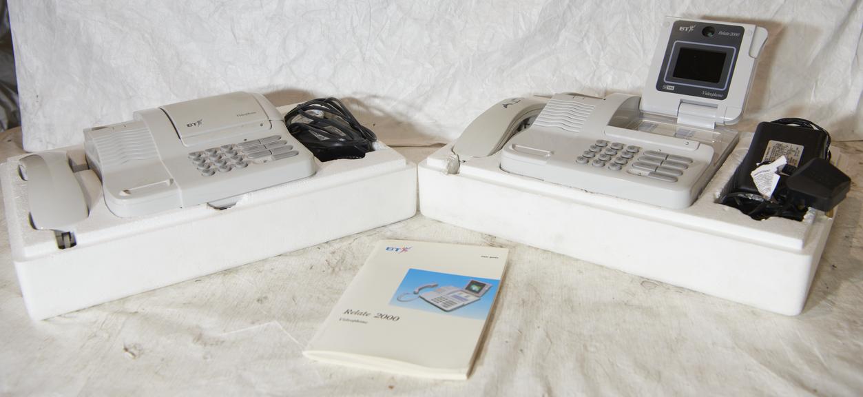 Two cream BT Relate 2000 videophones resting in white foam packaging, with one screen raised and the other folded down; handsets, coiled cables, power adapters and a Relate 2000 instruction booklet are arranged around them on a white cloth.