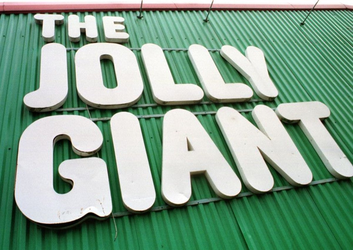 Close-up, upward-angled photograph of large white letters spelling “The Jolly Giant” mounted on a green corrugated building facade beneath red roof trim.
