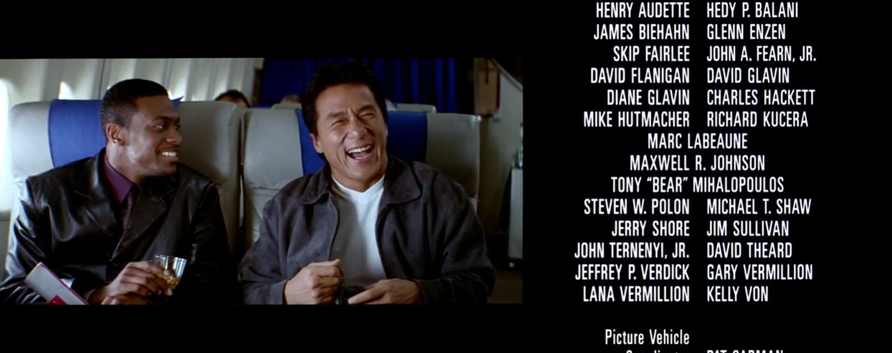 Two men seated beside each other in an aeroplane cabin smile and laugh in a video panel on the left, while columns of white production credits appear on black at right.
