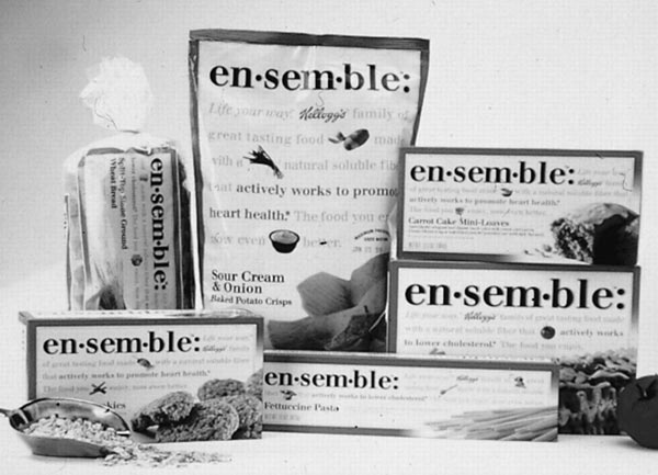 Black-and-white studio photograph of several en·sem·ble branded food packages arranged with plated food and ingredients. Visible labels include Sour Cream & Onion Baked Potato Crisps, Carrot Cake Mini-Loaves and Fettuccine Pasta.