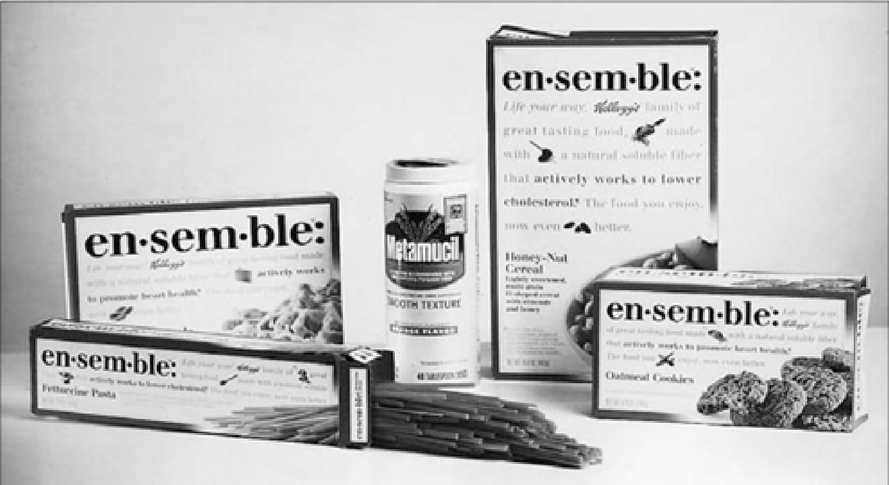 Black-and-white studio photograph of en·sem·ble packages, including several rectangular boxes, a tall cylindrical canister, and uncooked pasta laid across the foreground.