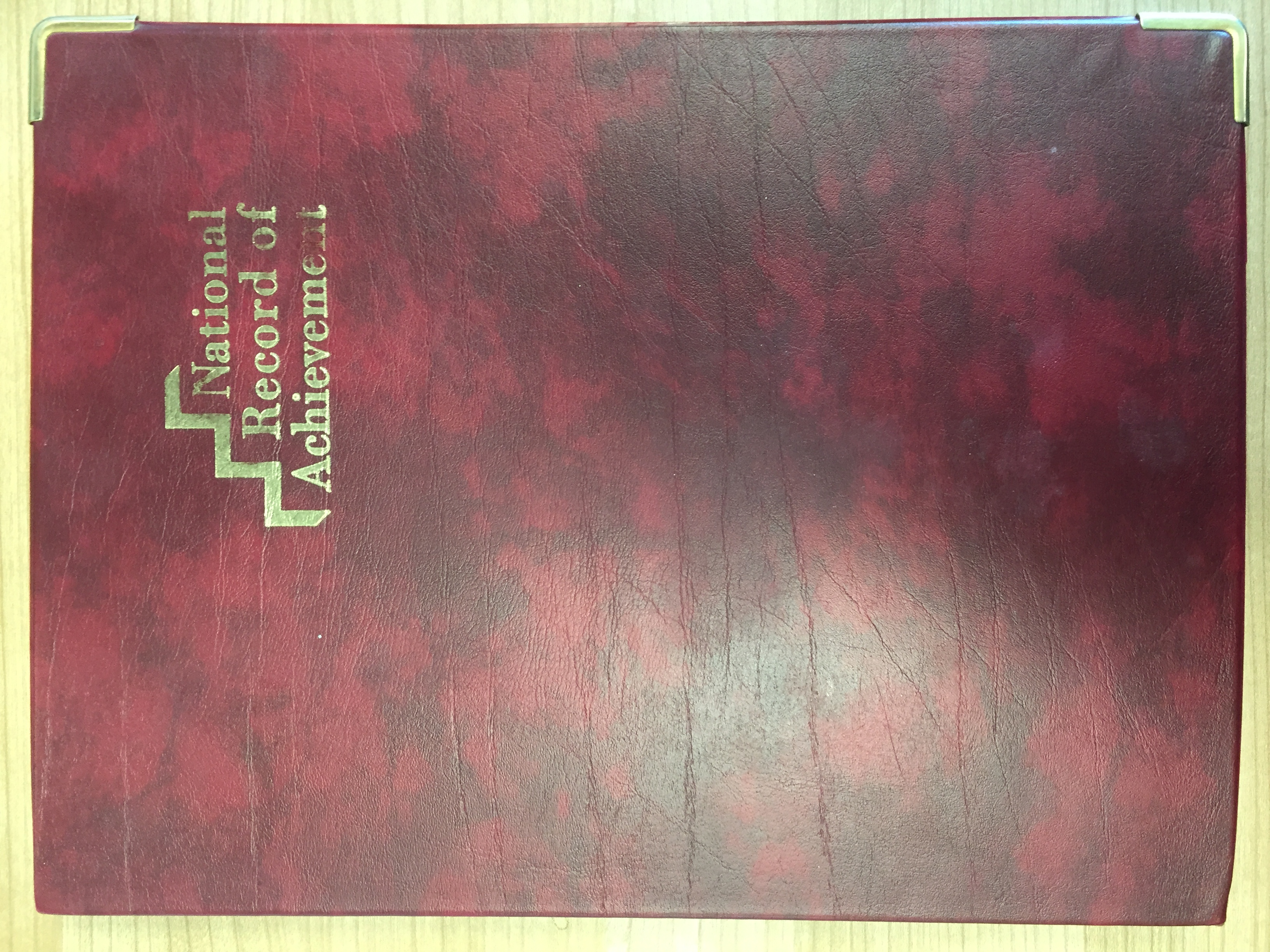 Closed burgundy, leather-textured folder on a pale wood surface, with a gold stair-step emblem and the words “National Record of Achievement” on the front. Silver-coloured protectors are fitted to the two visible right-hand corners, and the cover shows creases, scratches and reflected light.