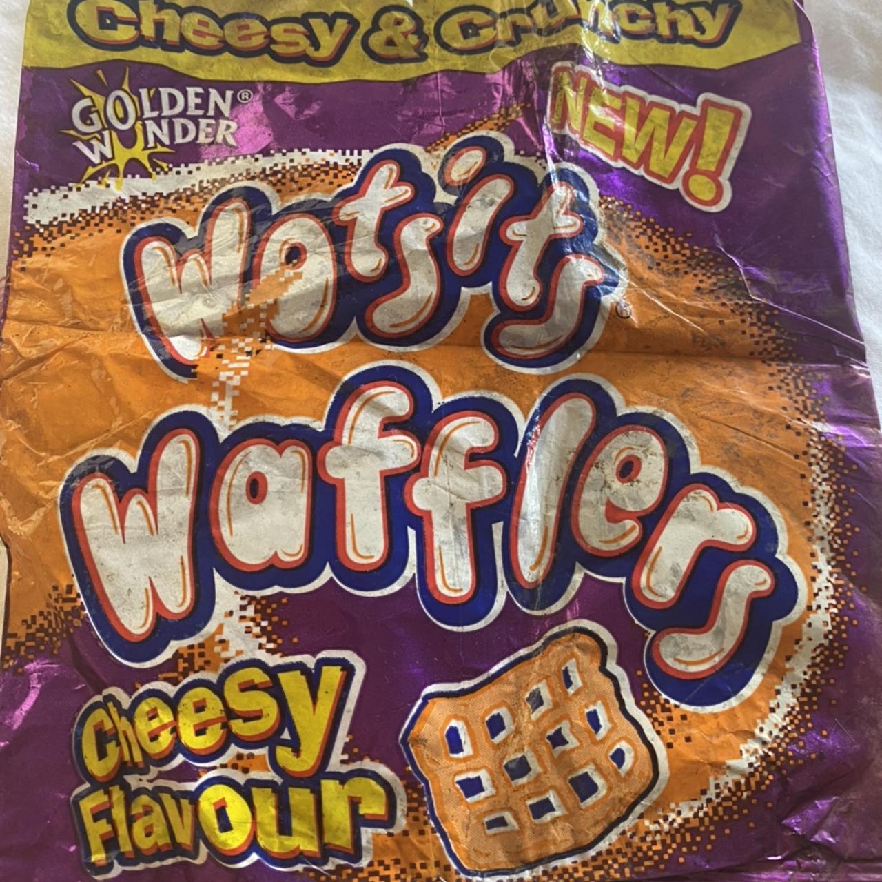 Close-up of a crumpled purple, orange and yellow snack packet printed with “Golden Wonder,” “Wotsits Wafflers,” “Cheesy Flavour,” “Cheesy & Crunchy” and “NEW!”, plus an illustration of a square lattice-shaped snack.