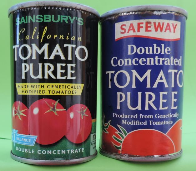 The black Sainsbury’s California Tomato Purée can stands beside the blue Safeway Double Concentrated Tomato Purée can. Both front labels explicitly identify genetically modified tomatoes.