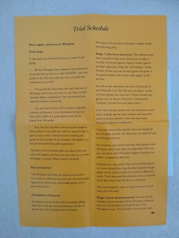 Photograph of an open yellow paper leaflet headed “Trial Schedule,” with two columns of printed text welcoming readers to Wireplay and describing setup, the trial period, features and development stages.