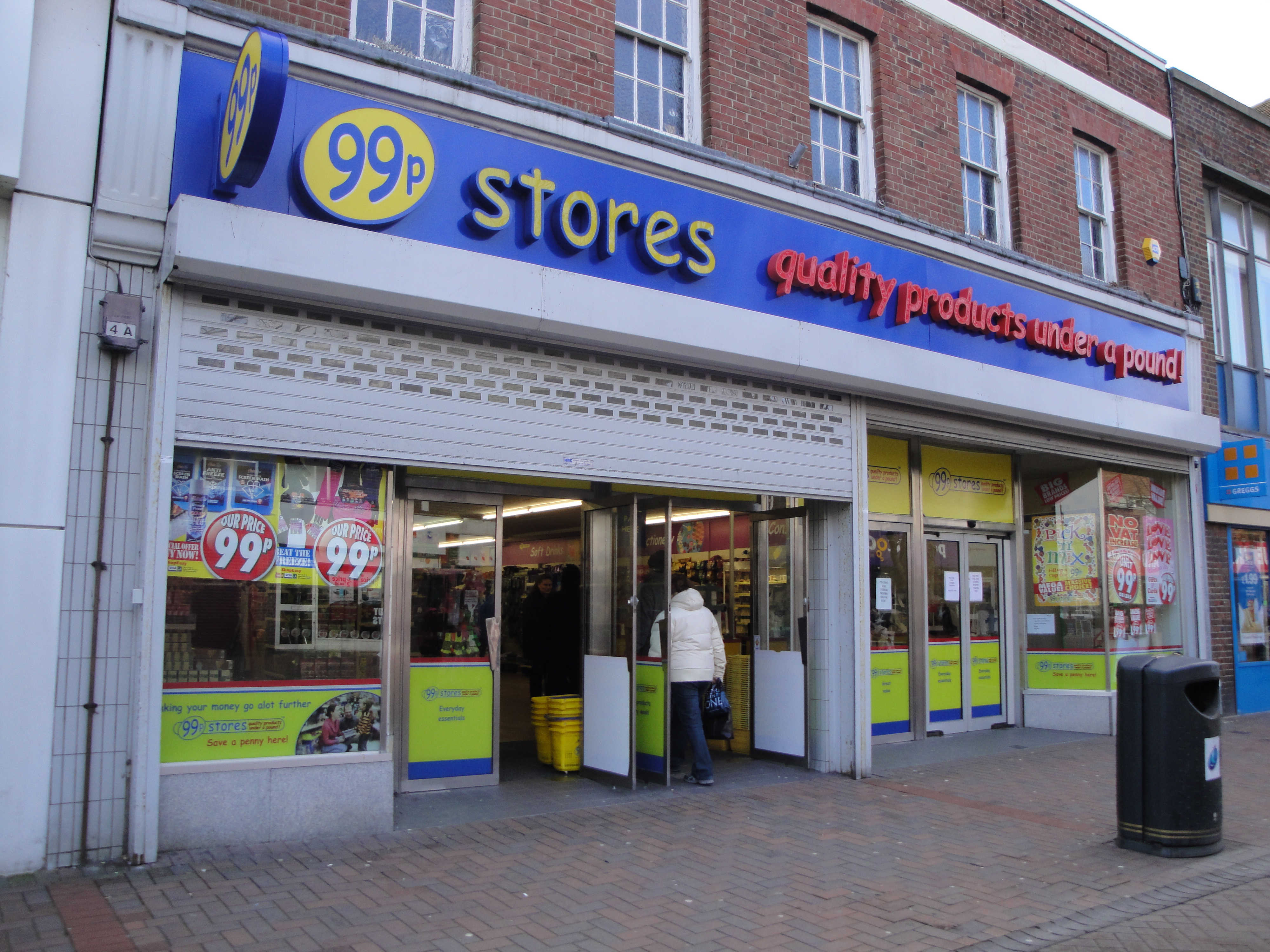A wide shopfront beneath a blue-and-yellow “99p Stores” sign and red lettering about products under a pound; circular window posters advertise “Our Price 99p,” and a person in a white hooded coat stands by the open entrance.
