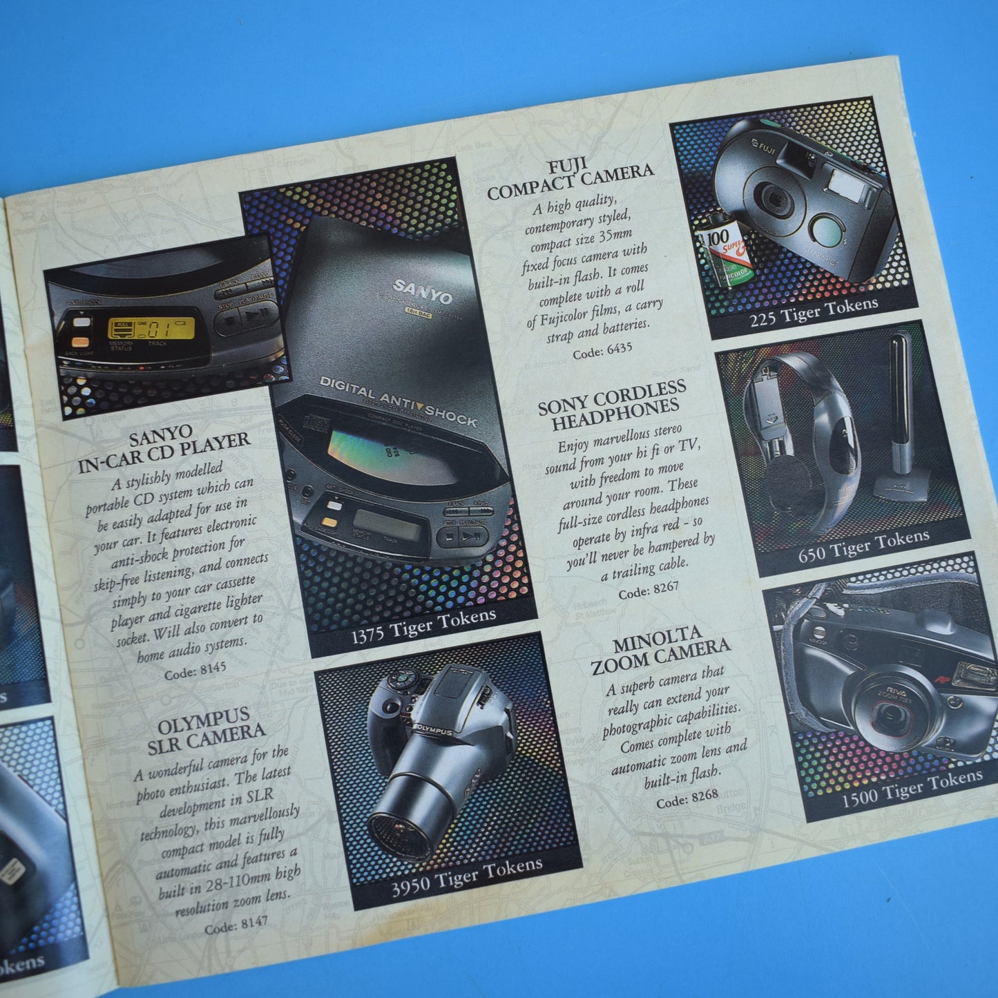 An open Esso Collection catalogue photographed on a blue surface, showing cameras, headphones and in-car audio equipment with descriptions and token prices.