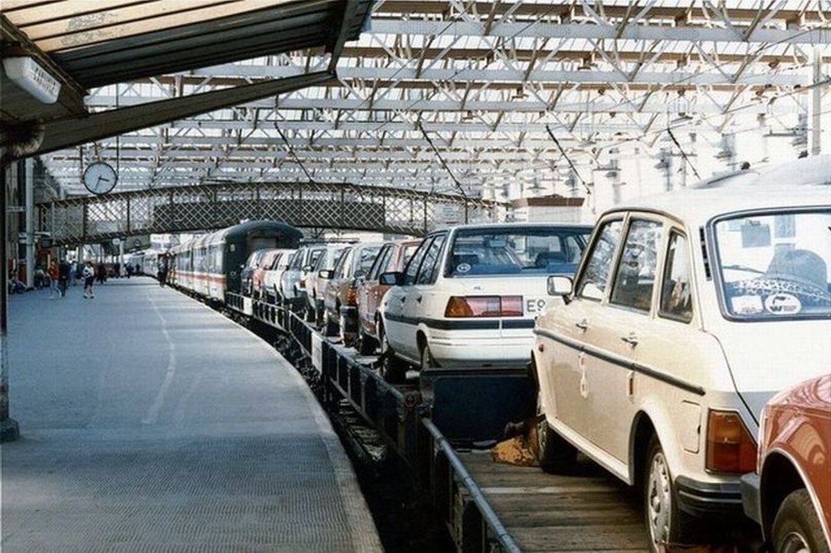 A row of cars loaded onto railway wagons beneath a large glazed station roof.