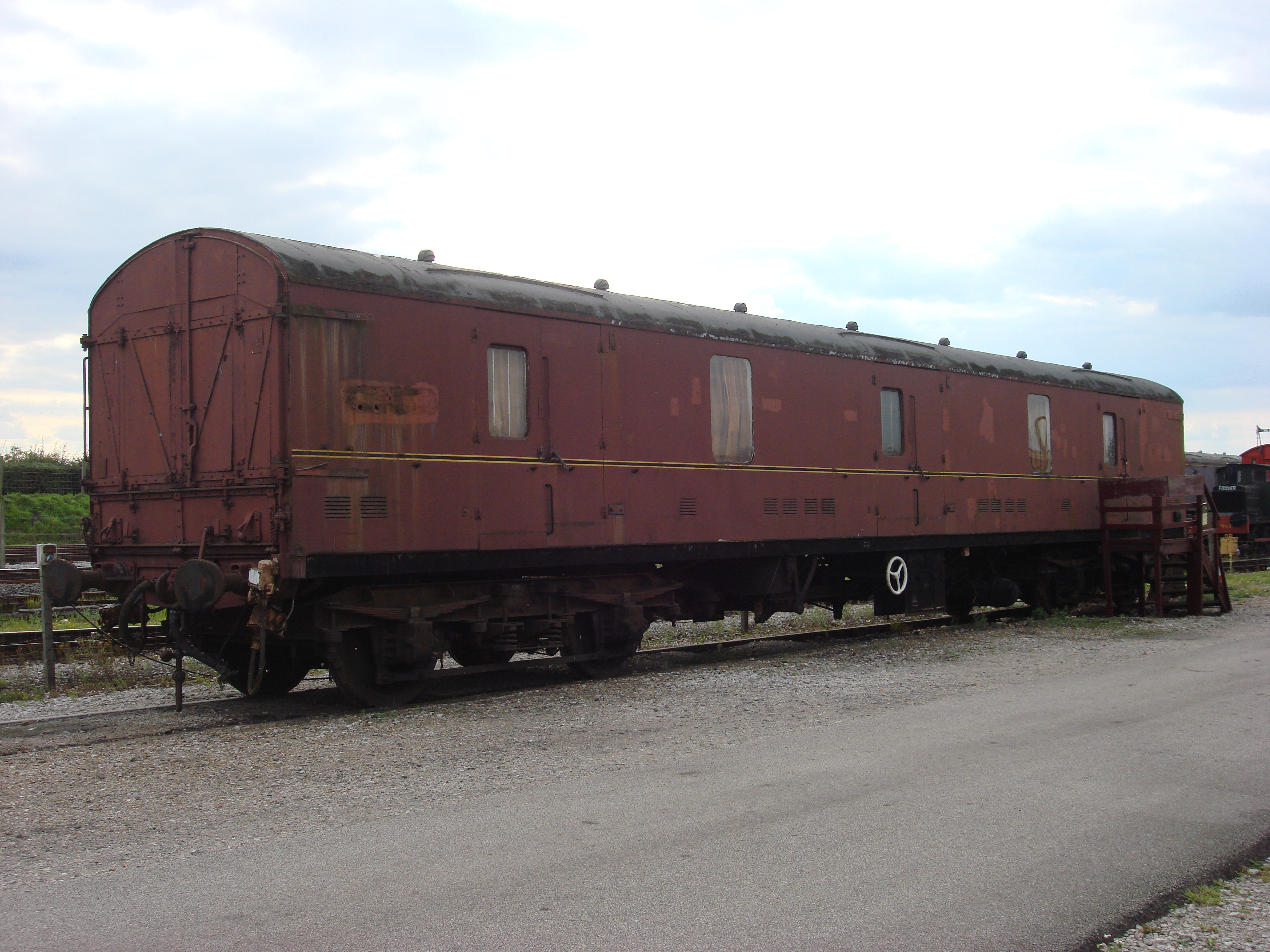 A preserved maroon railway van with large end doors and small windows stands outdoors beside railway tracks.