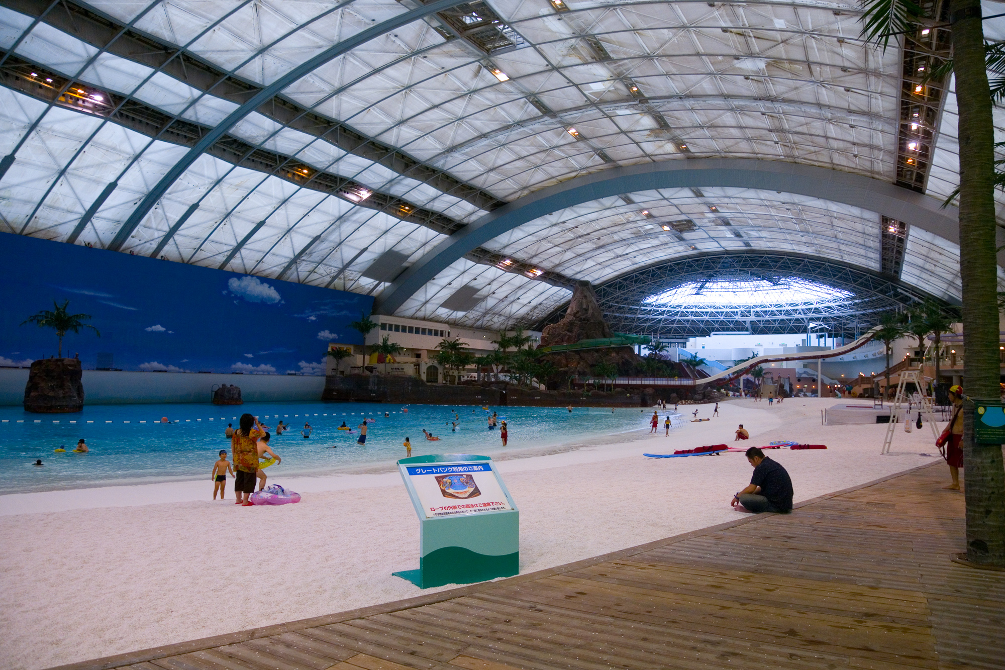 An indoor sandy beach and turquoise pool beneath a steel-framed roof, with painted clouds along the far wall.