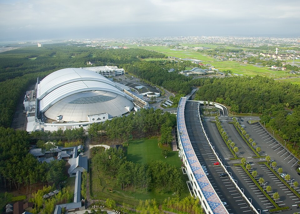 An enormous curved-roof building stands among trees beside a sweeping elevated road.
