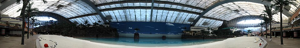 A wide panorama of an indoor beach and pool beneath arched roof beams, with palm trees at both sides.