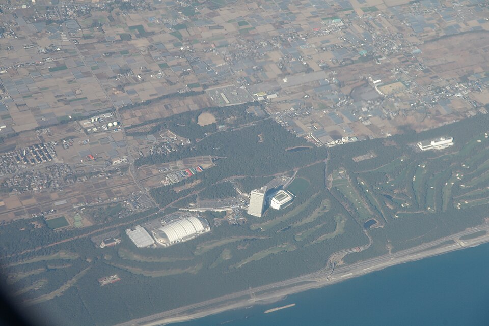 Aerial view of a coastal resort, with a long white-roofed building, hotel tower, forest belt and beach.