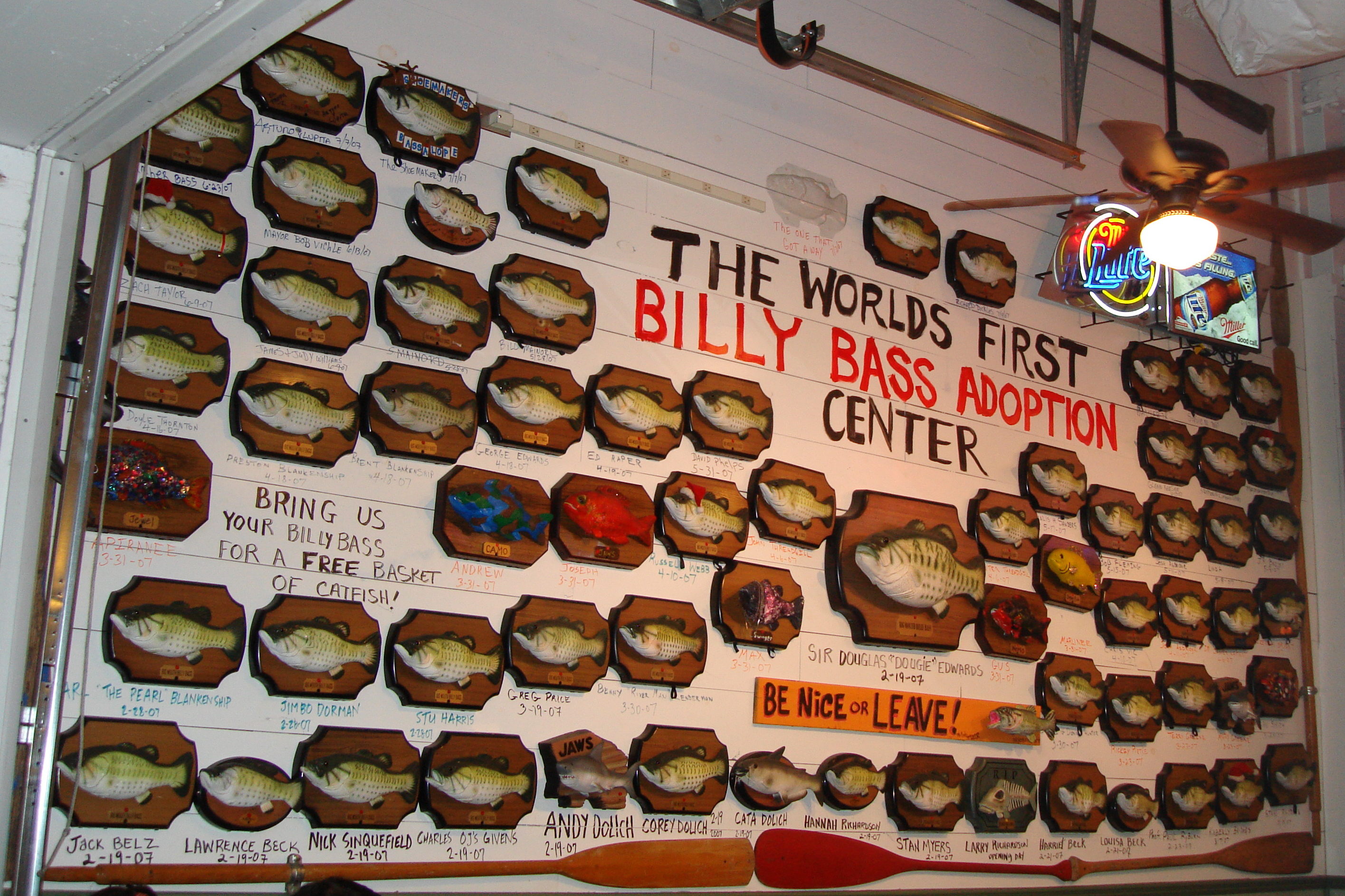 A wall crowded with dozens of mounted novelty fish on wood-grain plaques surrounds large hand-painted text reading “The World’s First Billy Bass Adoption Center.” Handwritten names and dates appear beneath many fish; a neon Miller sign, ceiling fan and wooden paddles are also visible.